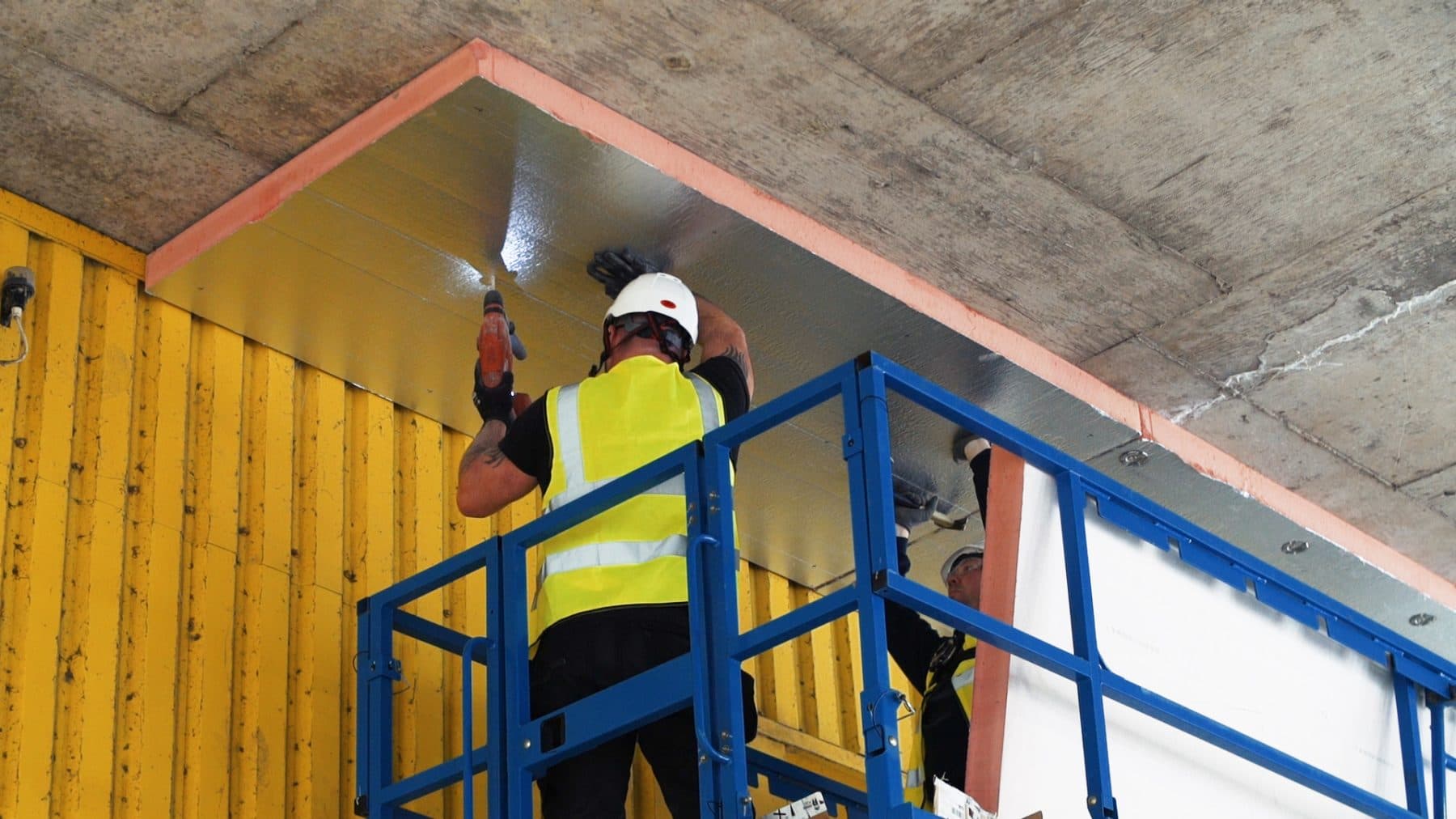 Crew installing commercial soffit insulation under an exposed undercroft from an elevated work platform